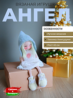 Вязаная кукла Ангел ручной работы в голубом капюшоне и белом платье сидит на картонной коробке на фоне новогодней елки.